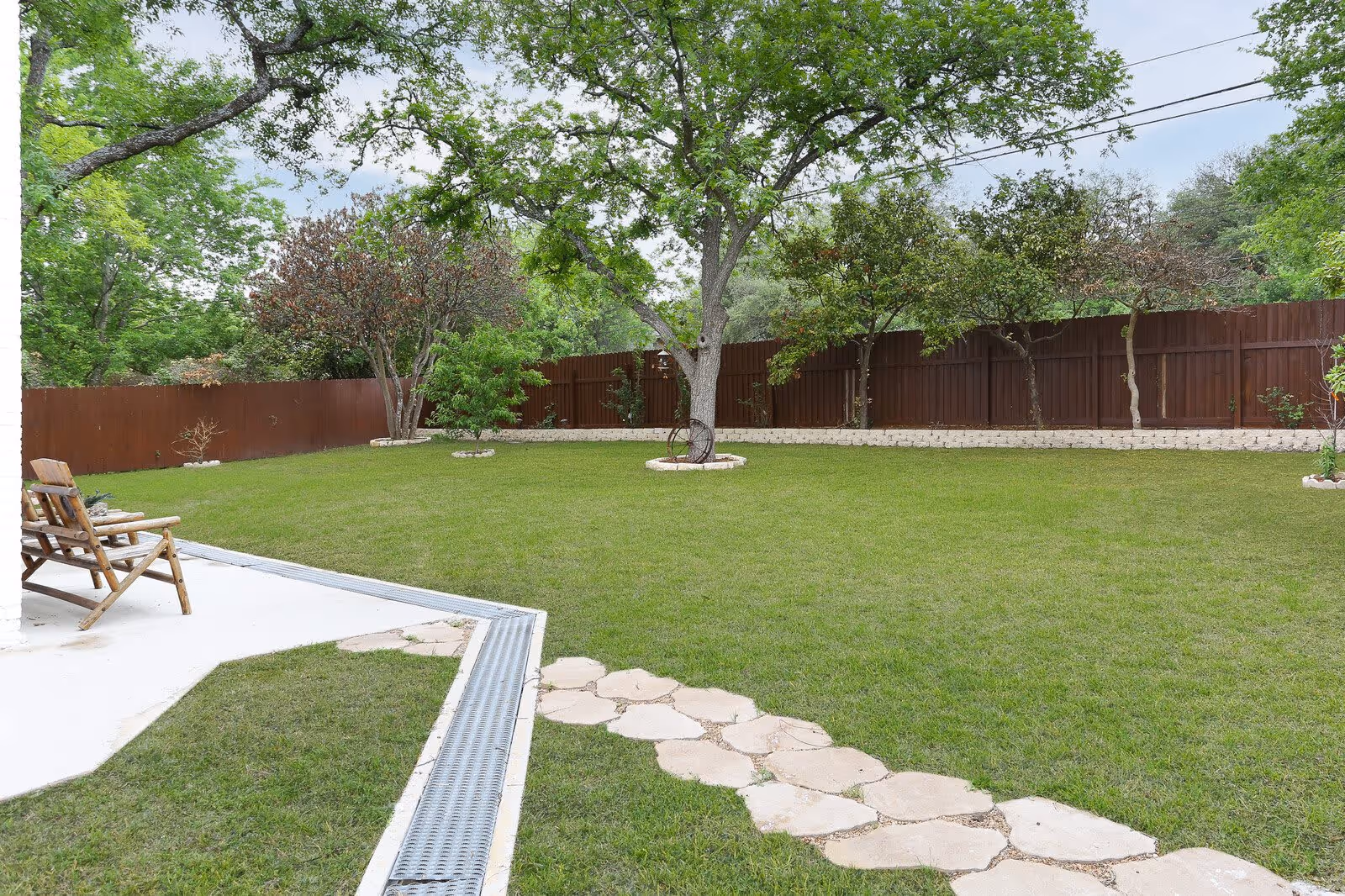 A spacious backyard with a well-maintained green lawn, a stone pathway, several trees, and a wooden fence surrounding the area. There is a wooden chair on a concrete patio on the left side of the image.