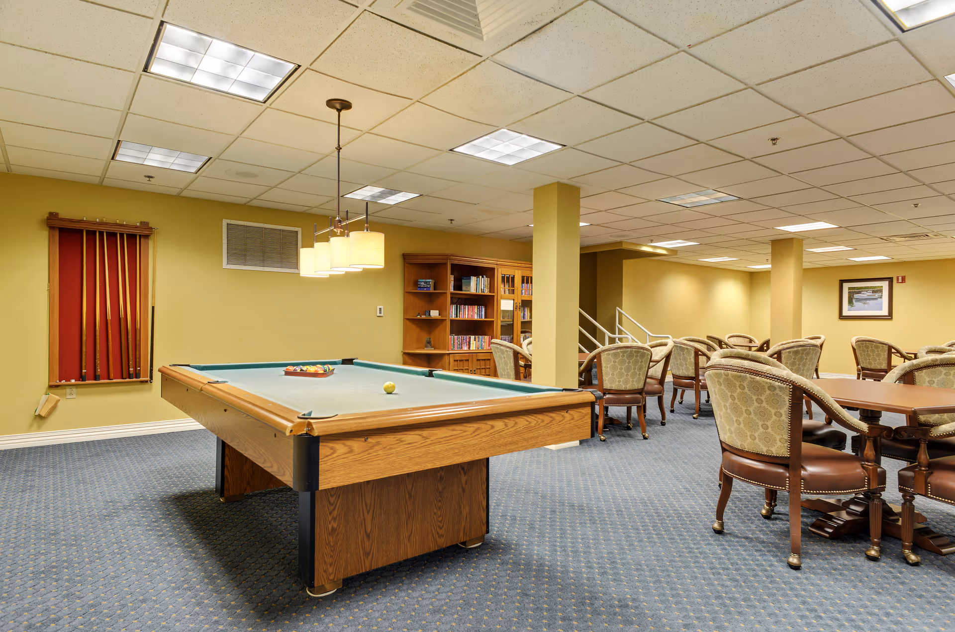 A recreational room featuring a pool table with billiard balls arranged on it, surrounded by several tables and upholstered chairs. The room has a blue carpet with a subtle pattern, yellow walls, and a ceiling with fluorescent lights. There is a wooden bookshelf filled with books against the wall and a rack holding pool cues.