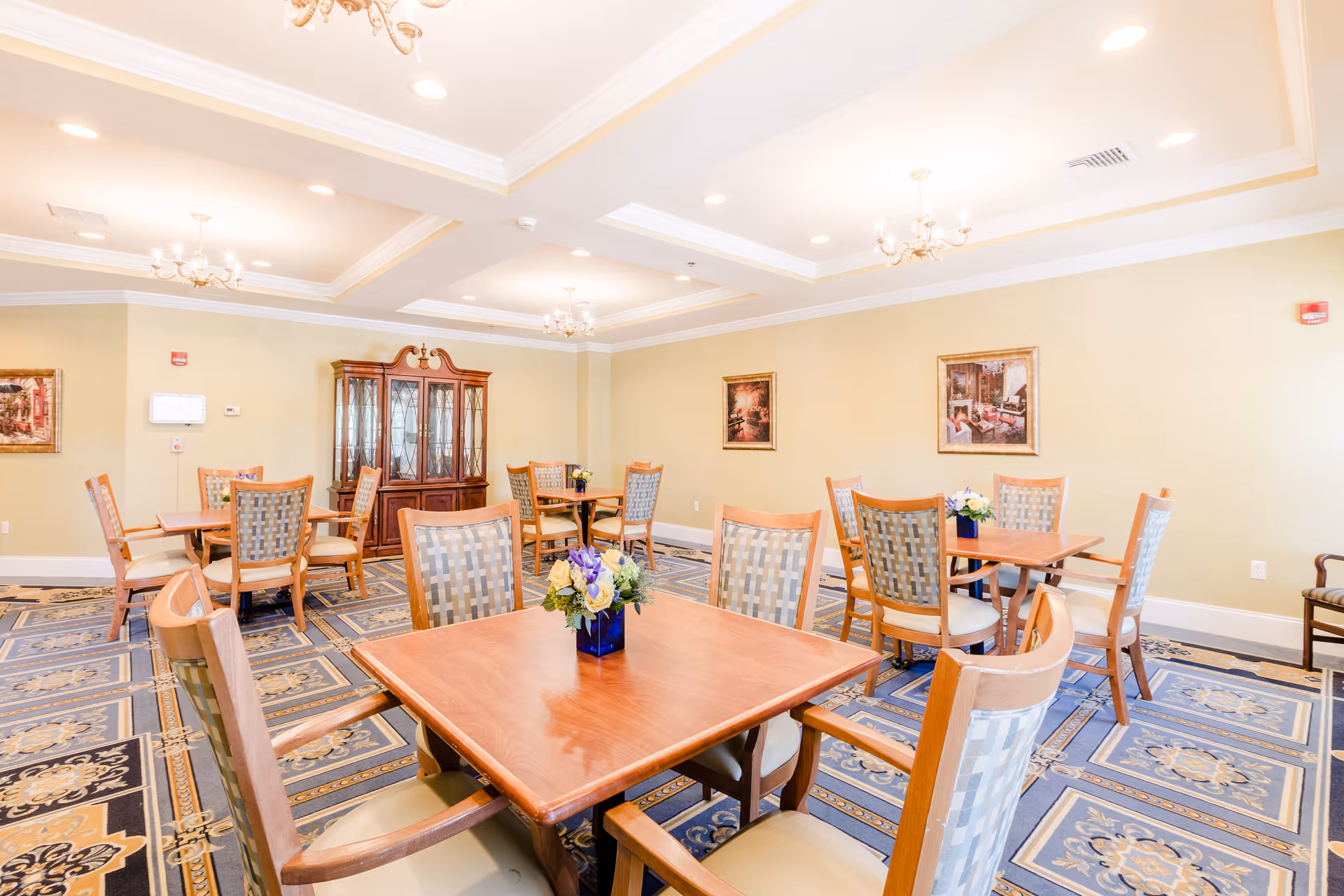 A bright and elegant dining room with several wooden tables and cushioned chairs arranged neatly. The room features a patterned carpet, chandeliers hanging from a coffered ceiling, framed artwork on the walls, and a wooden china cabinet in the background. Each table has a small floral centerpiece.
