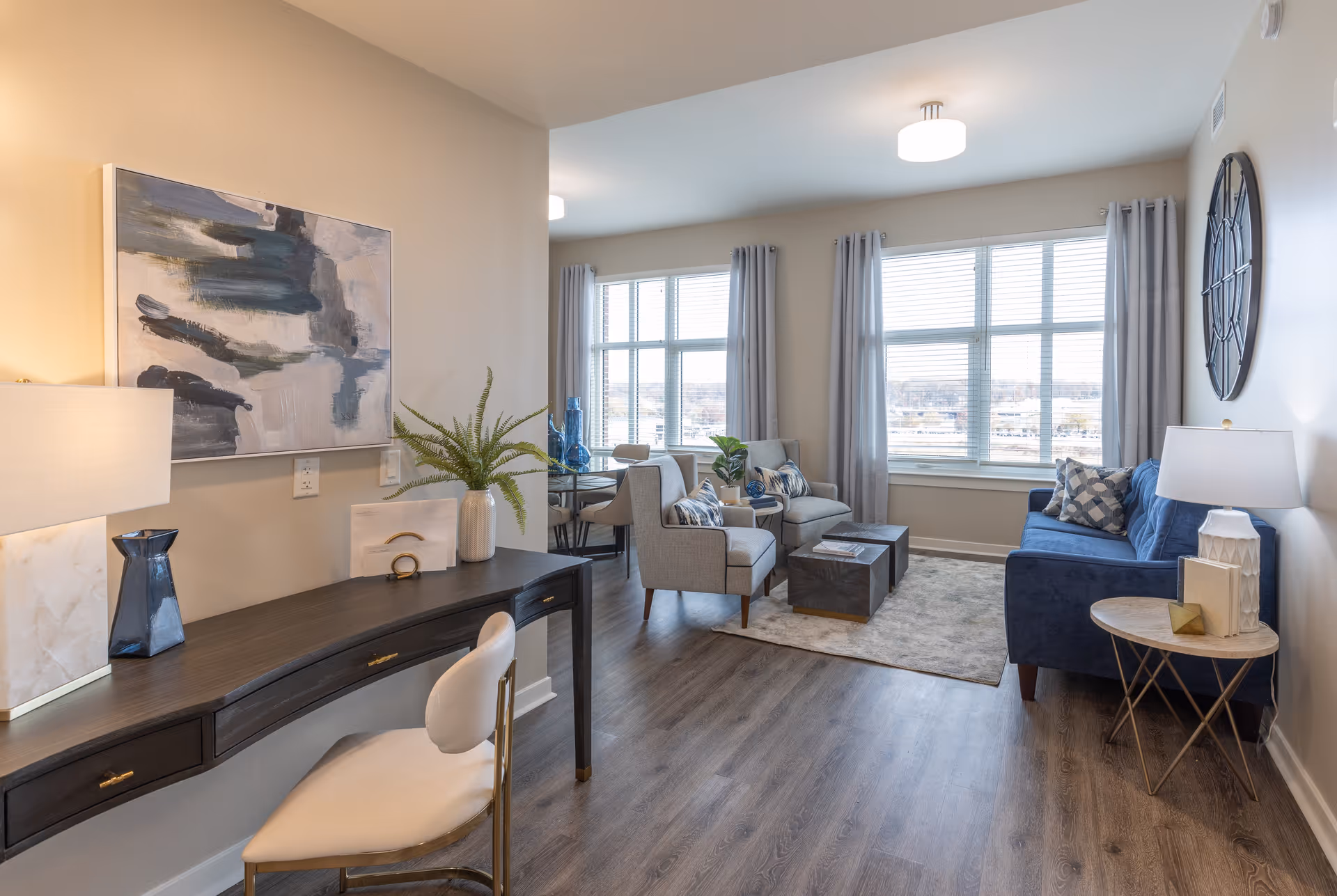 Bright, furnished living room with a desk in the foreground, blue sofa, armchairs and large windows letting in daylight.