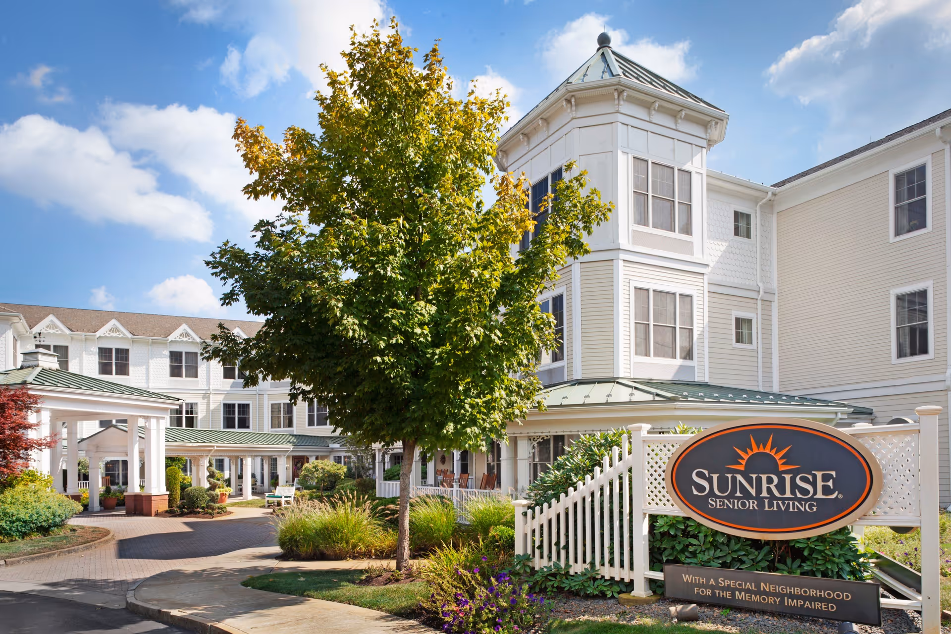 Exterior view of Sunrise Senior Living facility with a white multi-story building, green roofs, a large tree, landscaped garden, and a sign that reads 'Sunrise Senior Living with a special neighborhood for the memory impaired' under a blue sky with some clouds.