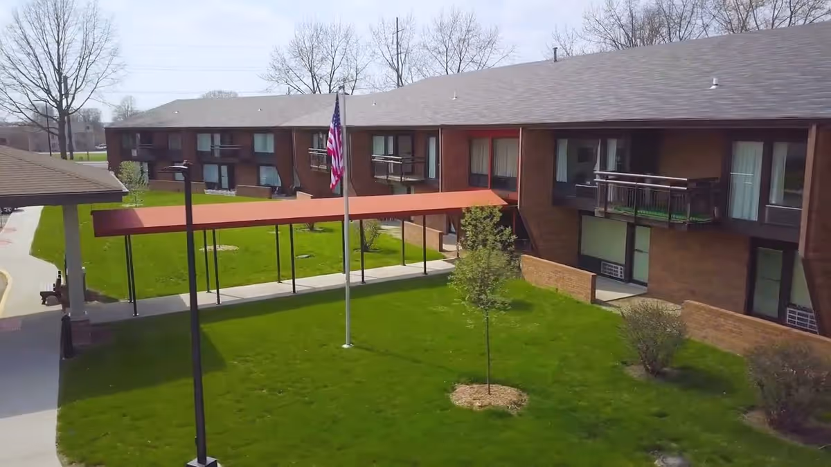Two-story brick senior living building with balconies surrounding a grassy courtyard, a covered walkway, and an American flagpole.