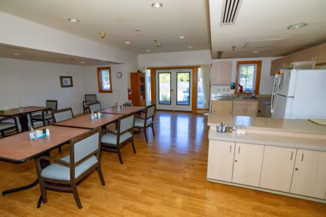 A bright communal dining area with wooden tables and chairs, a kitchenette along the right, and double glass doors at the back.