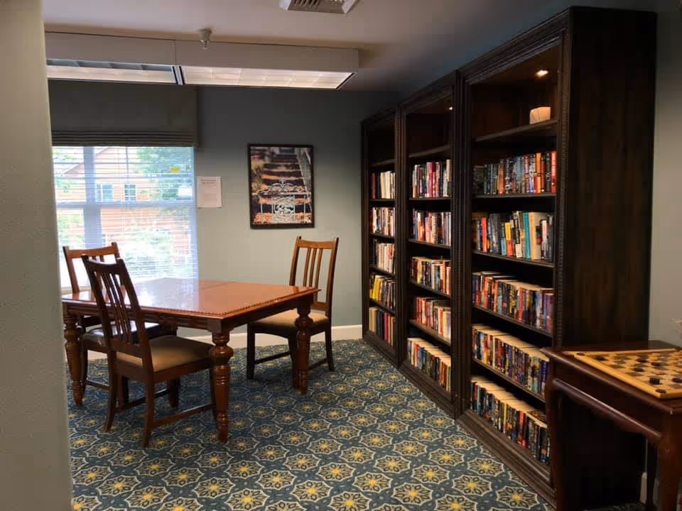 A cozy reading room with a wooden table and three chairs, a large window with blinds letting in natural light, a wall-mounted picture, and three tall bookshelves filled with books. The room has patterned carpet flooring and a small table with a checkerboard game on the right side.