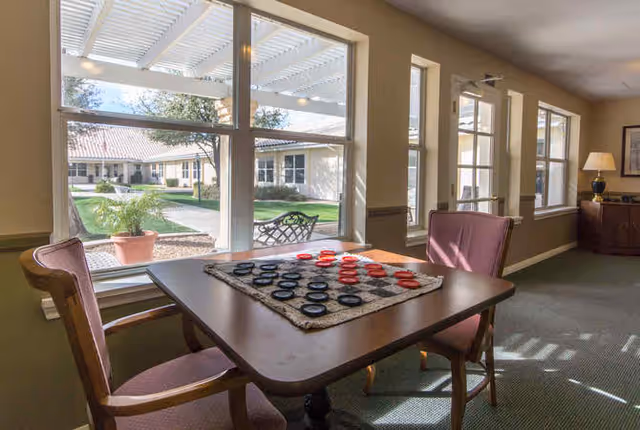 A cozy indoor seating area with a wooden table and two upholstered chairs. On the table is a checkerboard with red and black checkers set up for a game. Large windows behind the table provide a view of a sunny outdoor courtyard with a pergola, potted plants, and a bench. The room has beige walls, carpeted flooring, and a lamp on a side table in the background.