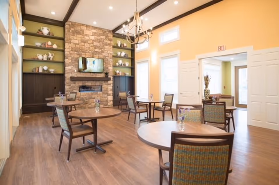 Bright communal dining room with round wooden tables and chairs, a stone fireplace with a mounted TV and built-in shelves.