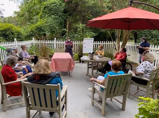 A group of elderly people sitting in a circle outdoors on a patio with chairs and tables, some under a red umbrella, listening to a person standing near a whiteboard. The area is surrounded by greenery and a white picket fence.