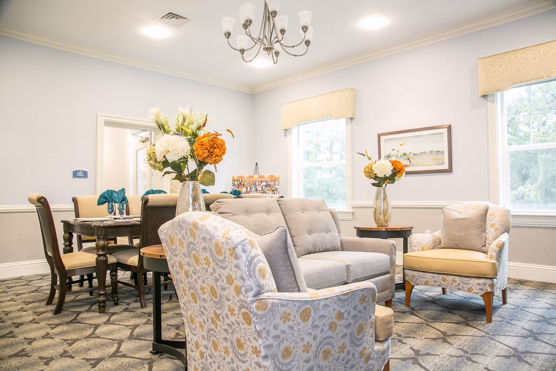A bright and cozy living room area in a senior living facility with a gray tufted sofa, two patterned armchairs with yellow cushions, a round wooden side table with a vase of flowers, and a dining table with chairs in the background. The room has light-colored walls, two windows with beige valances, a chandelier, and a framed painting on the wall.
