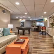 A spacious common area with a beige sofa and blue patterned pillows, an orange coffee table with books and a chessboard, a pool table in the center, and a bar counter with stools near a window. The room has wood-look tile flooring and recessed ceiling lights.