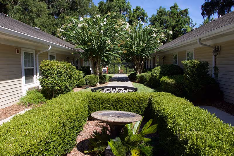 A well-maintained outdoor garden area between two beige buildings with white trim. The garden features neatly trimmed hedges, a stone birdbath, and a black metal bench along a paved walkway. Trees and bushes line the path under a clear blue sky.
