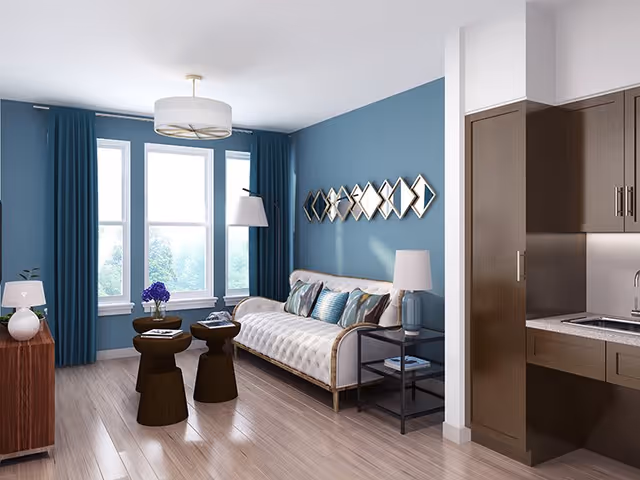 A modern living room with a white tufted sofa adorned with decorative pillows against a blue accent wall. Above the sofa is a geometric mirror art piece. The room features three large windows with blue curtains, two dark wooden side tables with a vase of purple flowers and books, a floor lamp, and a side table with a white lamp. To the right, part of a kitchenette with brown cabinets and a sink is visible. The floor is light wood.