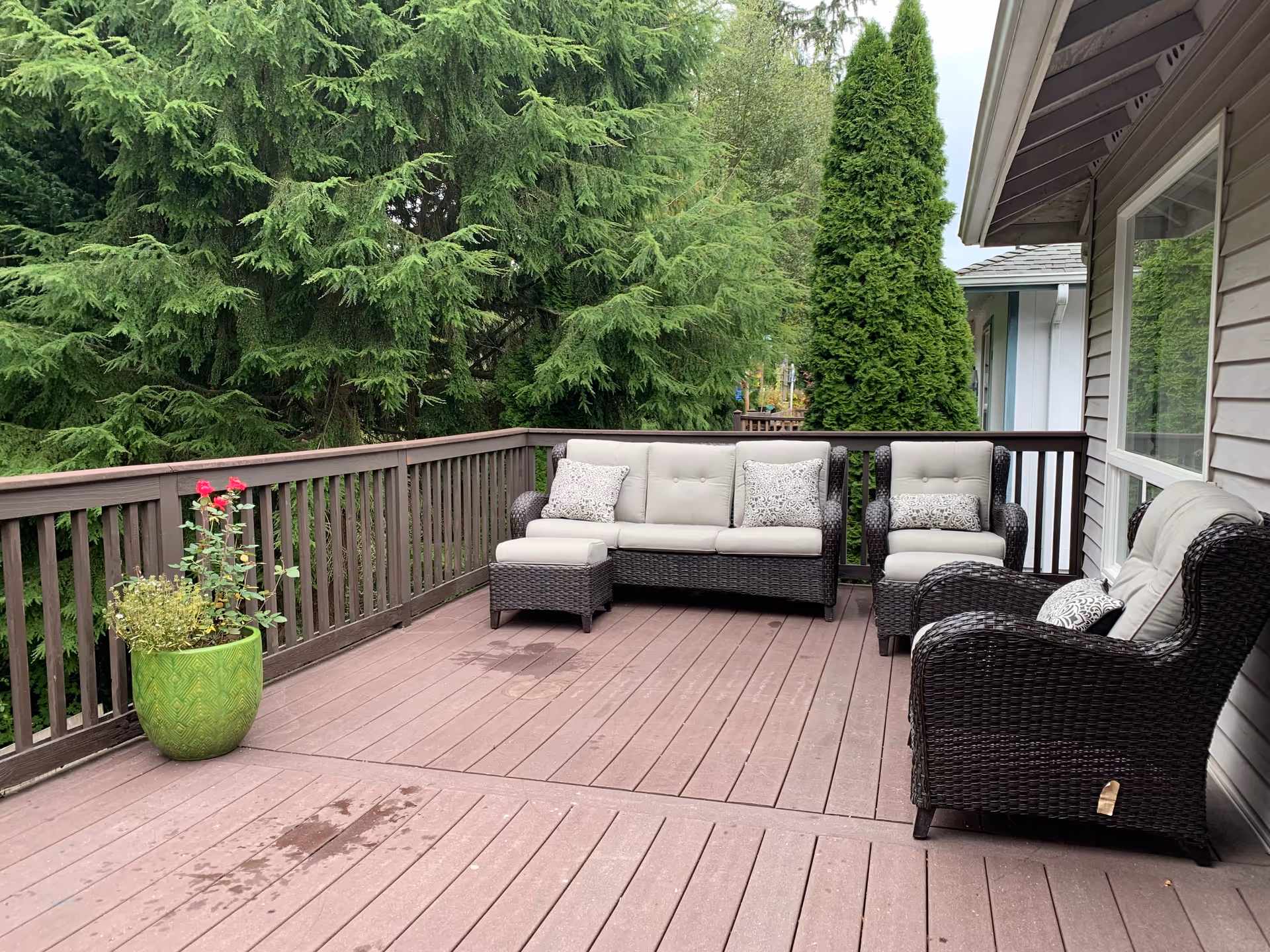 Outdoor wooden deck with dark brown wicker patio furniture including a sofa, two armchairs, and an ottoman with light gray cushions and patterned pillows. A green potted plant with red flowers is placed near the wooden railing. Tall green trees and shrubs surround the deck, and part of the building with beige siding and a window is visible on the right.