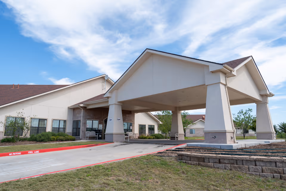Exterior view of Las Alturas Nursing & Transitional Care facility showing a covered entrance with large pillars, a driveway with red painted curbs marked 'No Parking Fire Lane', and a well-maintained lawn and shrubs under a partly cloudy blue sky.