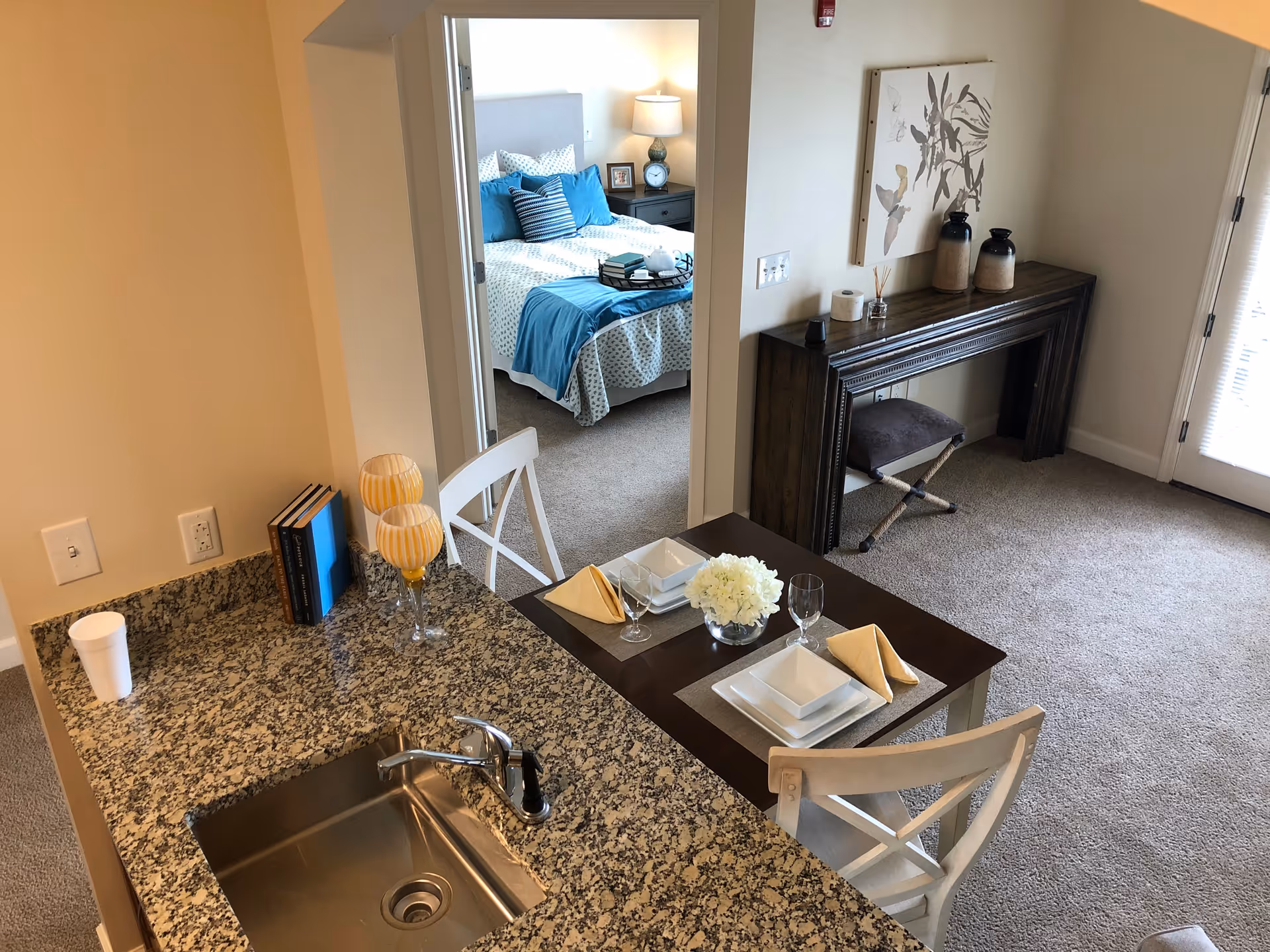 View of a small dining area with a table set for two, adjacent to a kitchen counter with a sink. In the background, there is a bedroom with a made bed featuring blue and white bedding. The living area has a decorative console table with vases and a piece of wall art above it. The room is carpeted and well-lit with natural light coming from a door with blinds.