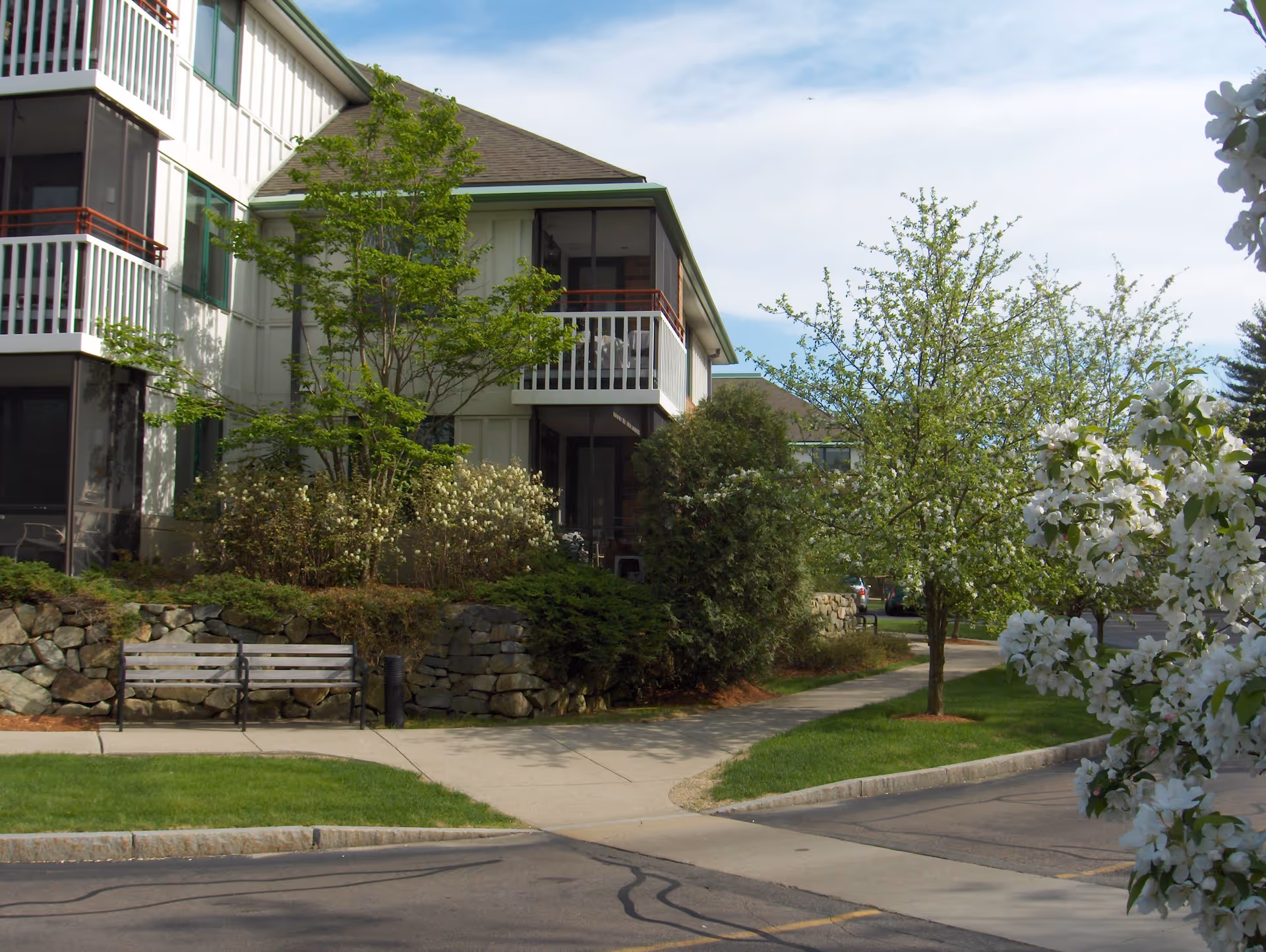 Exterior view of a senior living facility building with white siding and balconies, surrounded by green trees, bushes, and blooming white flowers. A stone retaining wall and a wooden bench are visible near a sidewalk leading to the entrance.
