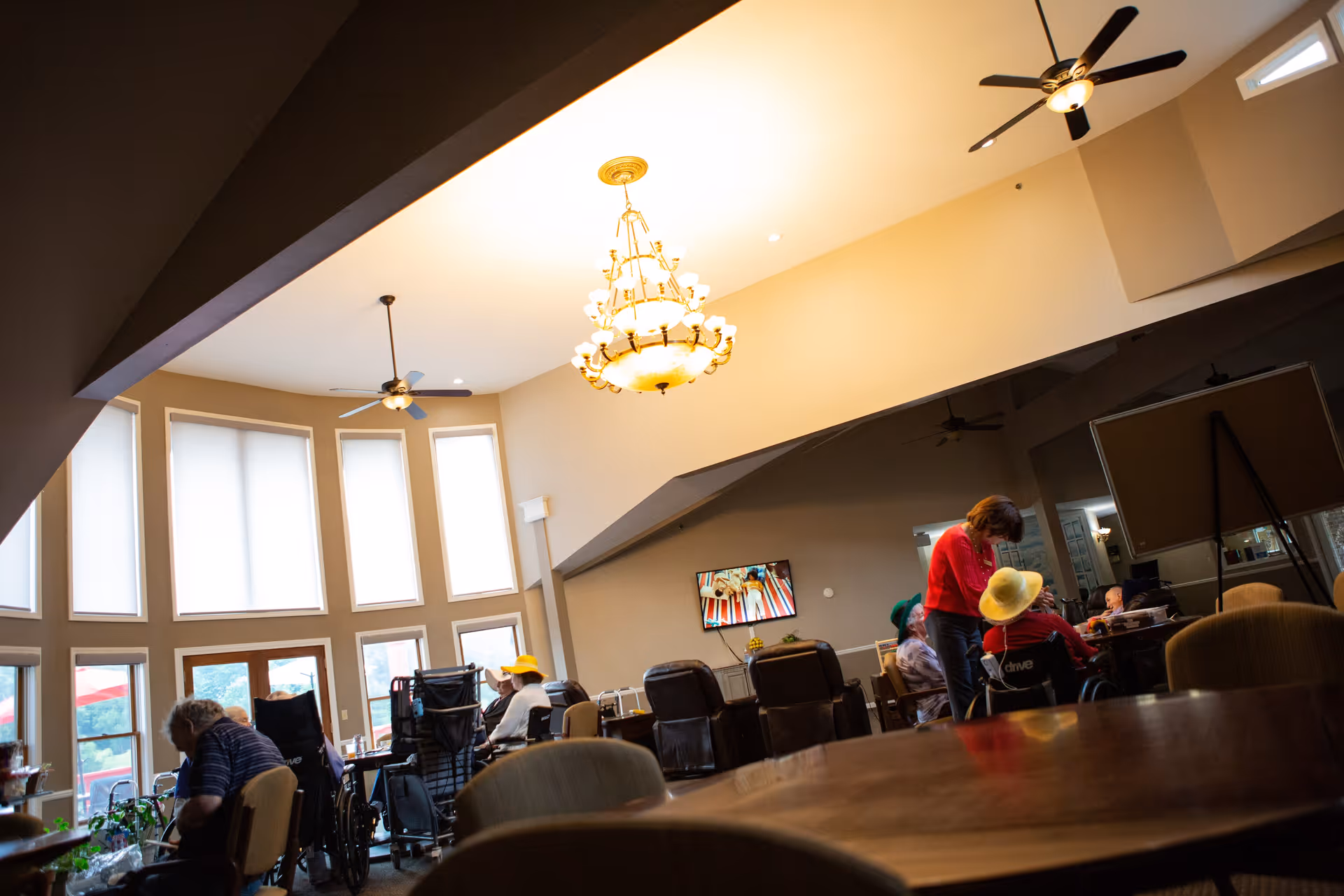 A spacious common area in a senior living facility with large windows, ceiling fans, and a chandelier. Several elderly residents, some in wheelchairs, are seated and engaged in activities. A caregiver in a red top is interacting with one of the residents wearing a yellow hat. A television is mounted on the wall in the background.