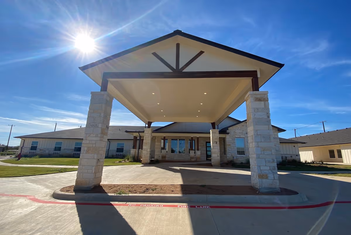 Front exterior view of Bella Groves facility showing a covered entrance with stone pillars under a clear blue sky with the sun shining brightly.
