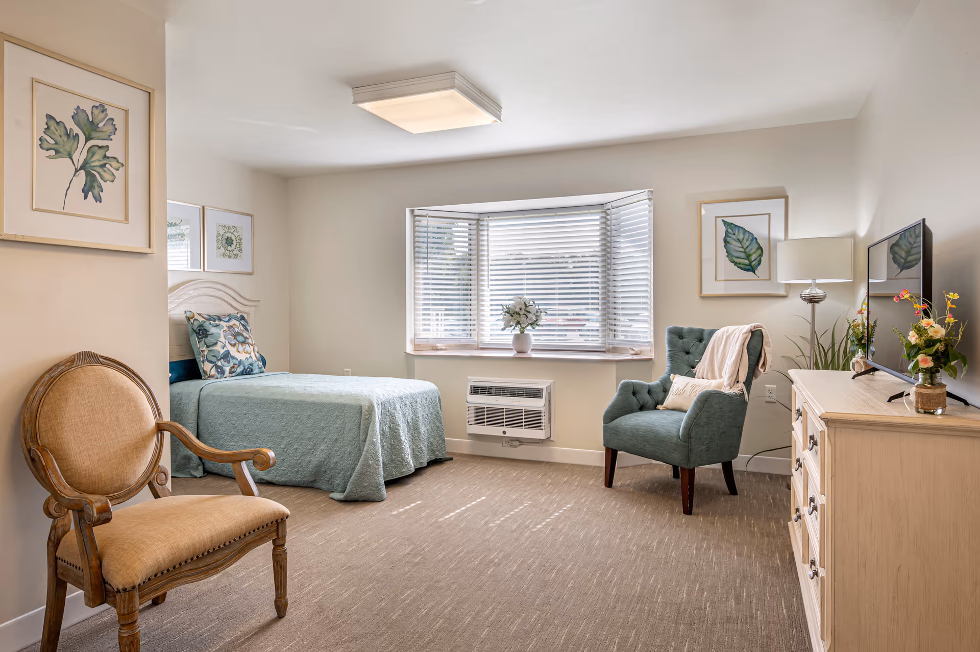 A bright and cozy bedroom in a senior living facility featuring a single bed with a light blue quilt and floral pillow, a wooden armchair with beige upholstery, a green cushioned armchair with a throw blanket and pillow, a wooden dresser with a flat-screen TV and flower vases, a bay window with white blinds, and framed botanical artwork on the walls.