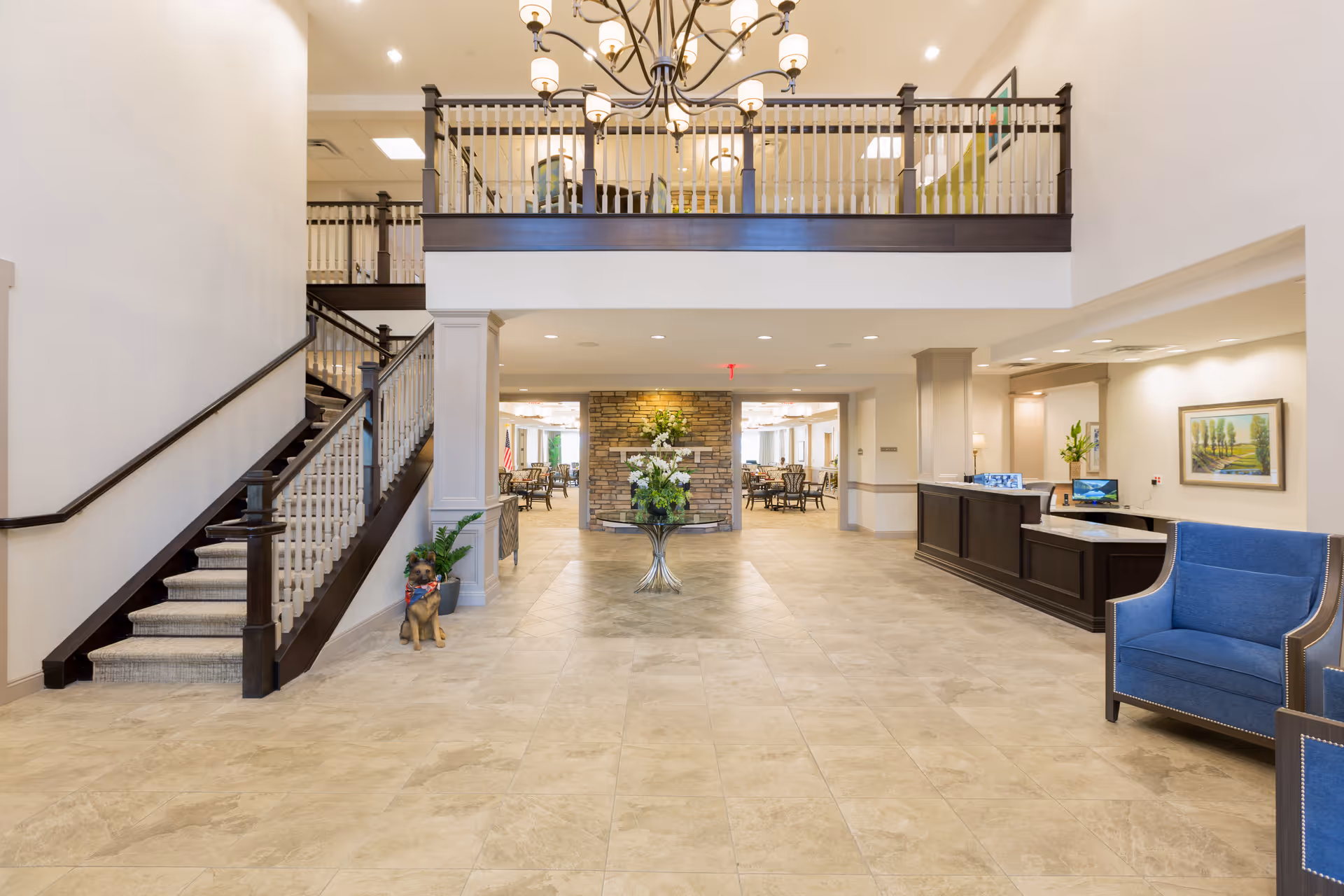 Spacious and well-lit senior living facility lobby with a tiled floor, a staircase with dark wood railing on the left, a reception desk on the right, and a blue armchair nearby. A decorative table with a floral arrangement is centered under a mezzanine with additional seating. The background shows a dining area with tables and chairs.
