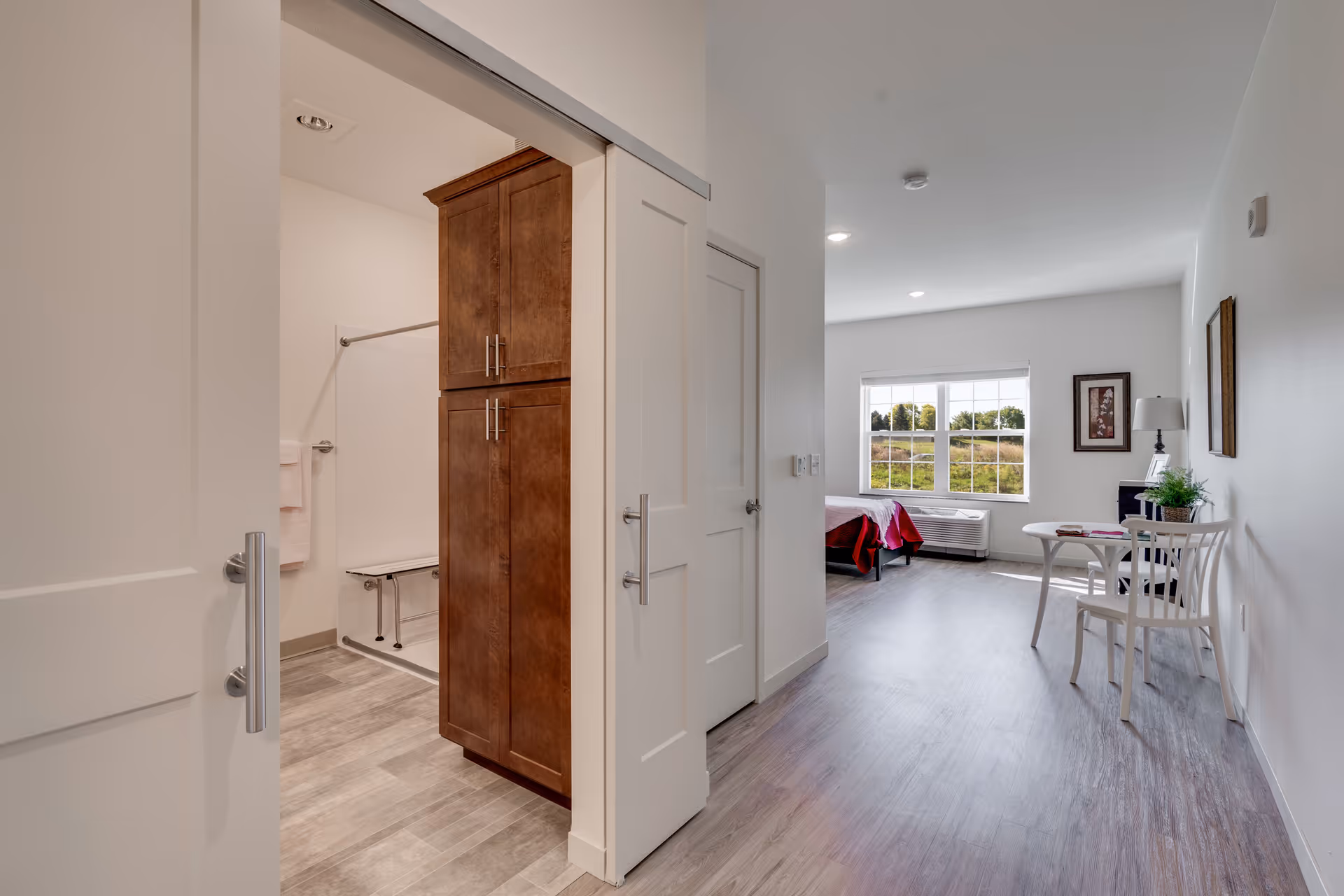 Interior view of a senior living facility room showing a hallway with a bathroom on the left featuring a wooden cabinet and a shower with a bench. The hallway leads to a bedroom area with a bed covered in a red blanket, a window with a view of greenery outside, a small white table with two chairs, a lamp, and framed artwork on the walls.
