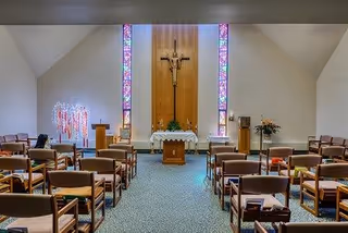 Interior view of a chapel with rows of wooden chairs facing an altar. The altar is adorned with a white cloth and a crucifix mounted on a wooden panel between two tall stained glass windows. There are floral arrangements on either side of the altar and a lectern to the left.