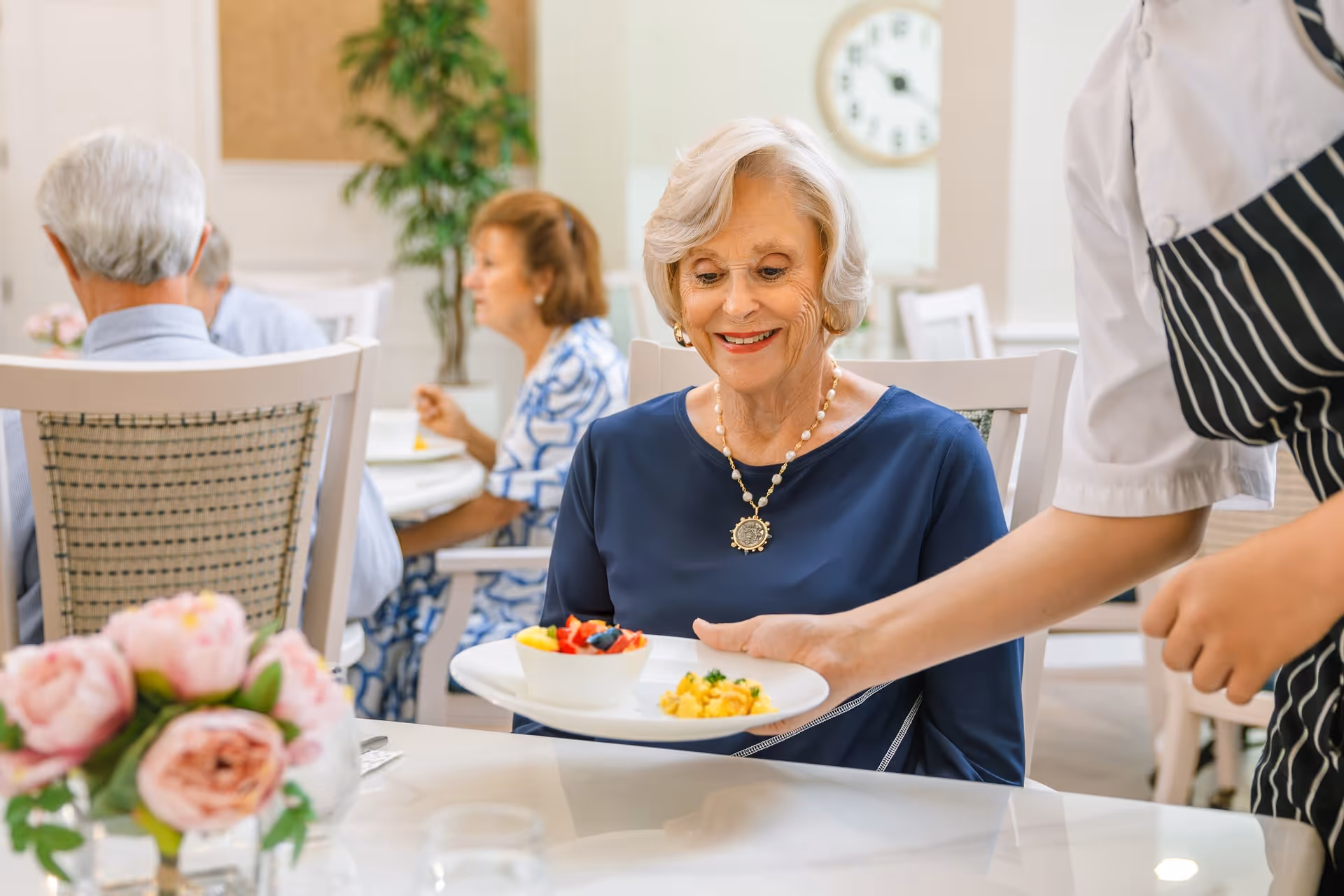 An elderly woman with short gray hair and a blue top is smiling as a server in a striped apron places a plate of food in front of her at a dining table. In the background, other elderly people are seated and eating in a bright, well-lit dining room with white chairs and a clock on the wall.
