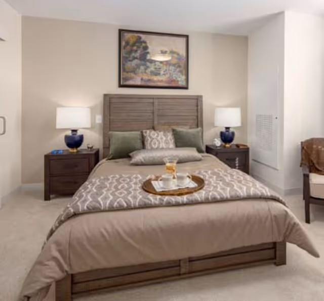 A neatly made bedroom with a wooden bed, matching nightstands and lamps, a framed painting above the headboard, and a breakfast tray on the bed.