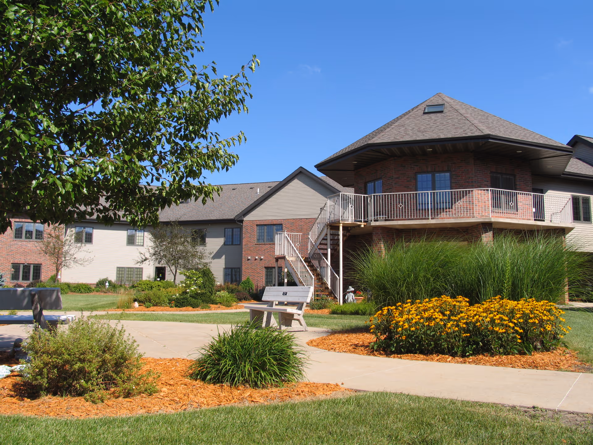Outdoor view of a senior living facility with a well-maintained garden featuring green shrubs, yellow flowers, and a tree. There is a concrete pathway with a bench along it, and a two-story building with brick and siding exterior in the background under a clear blue sky.