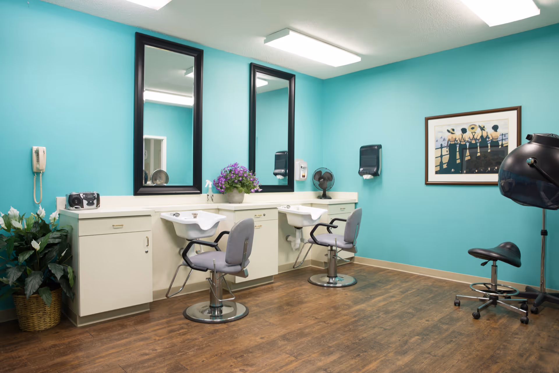An interior salon room with two styling chairs and washbasins, large mirrors, teal walls, a hooded hair dryer, and wood flooring.