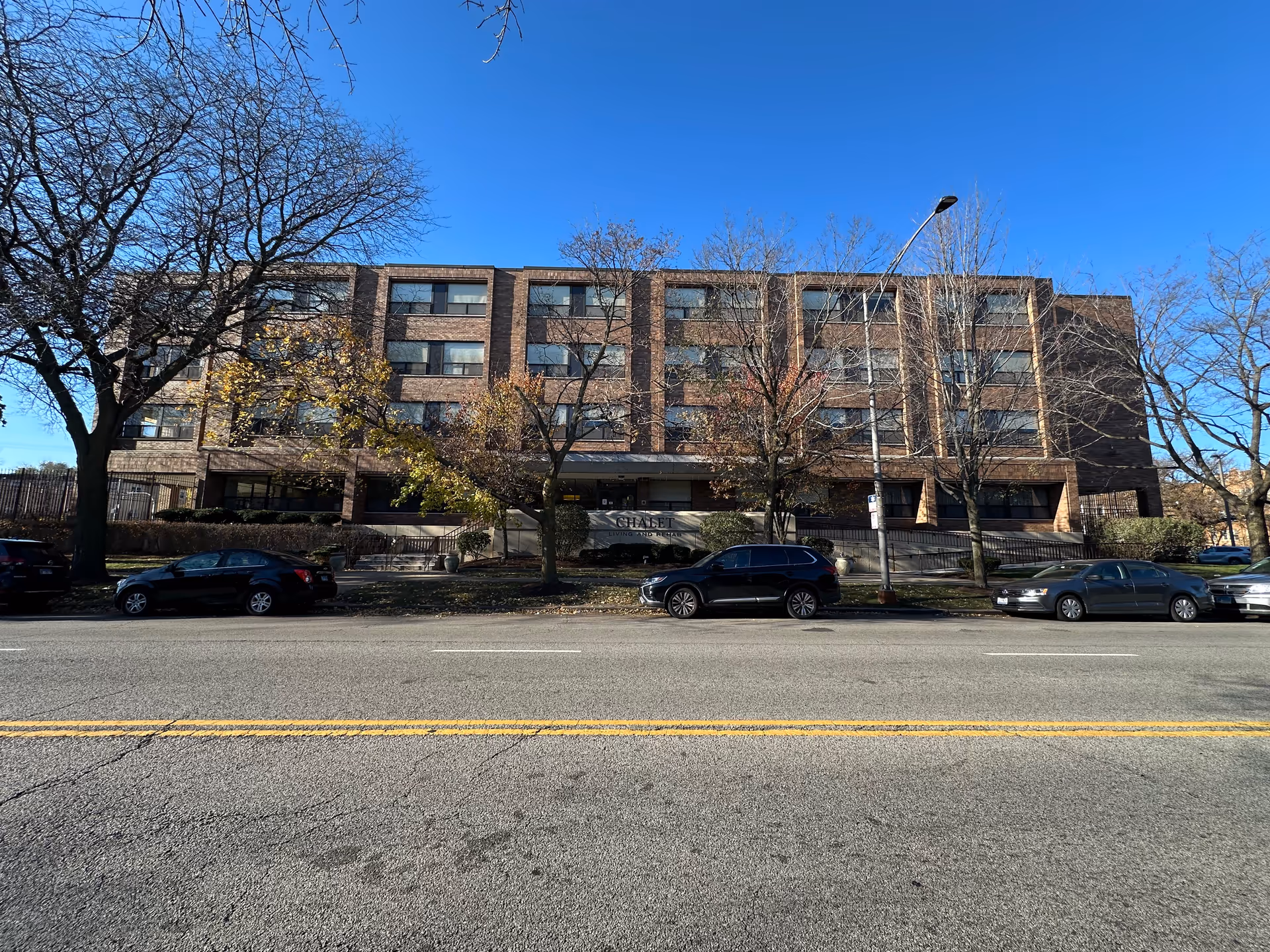 Exterior front view of a four-story brick building with several windows, trees with sparse leaves, parked cars along the street, and a clear blue sky above. A sign in front of the building reads 'CHALET'.