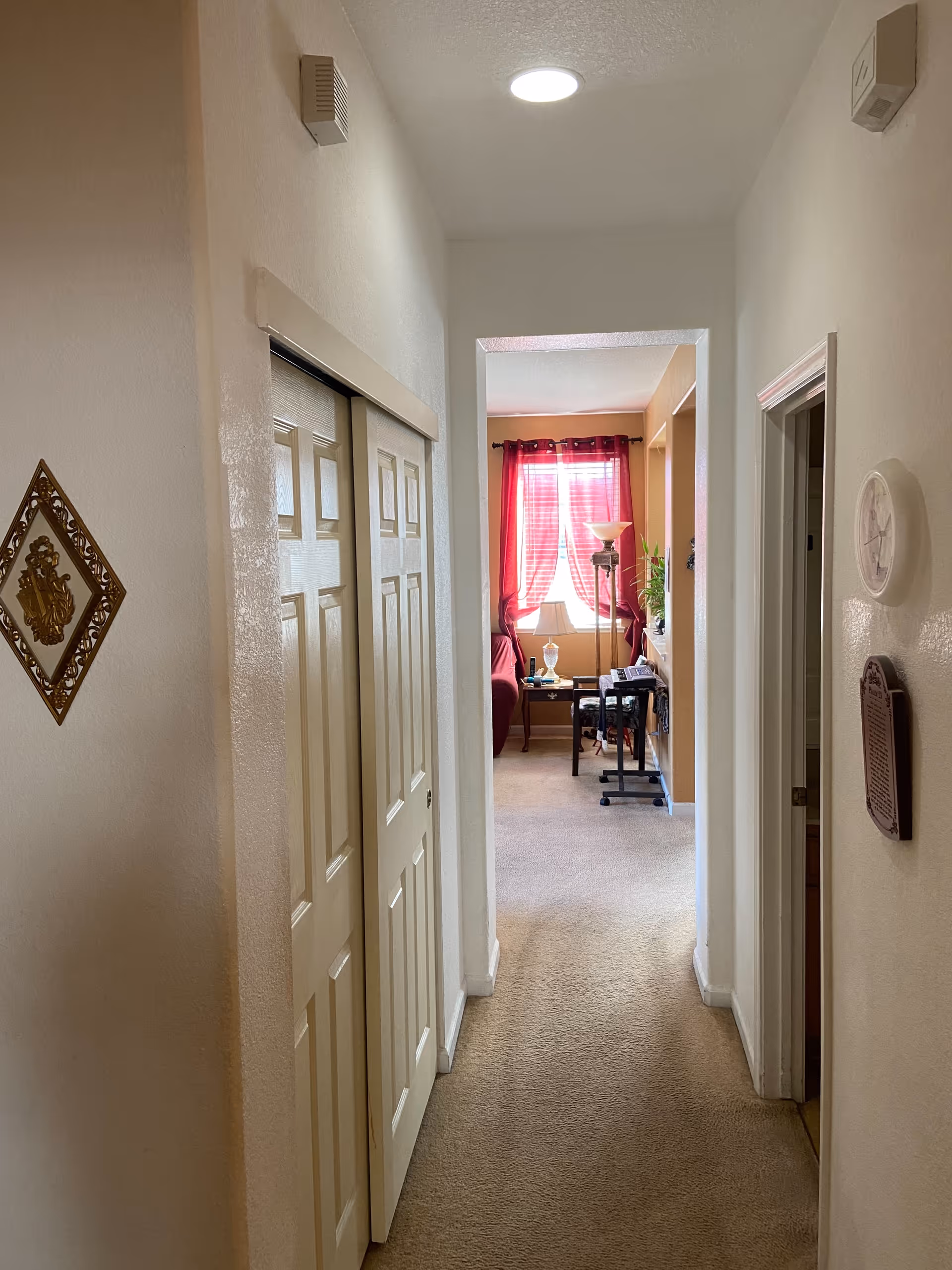 A narrow hallway with beige carpet and white walls leading to a living room area with a window covered by red curtains. On the left side of the hallway, there are two closed closet doors and a decorative wall hanging. On the right side, there is a doorway and a round wall clock. The living room at the end of the hallway has a floor lamp, a small table with a lamp, and a chair.