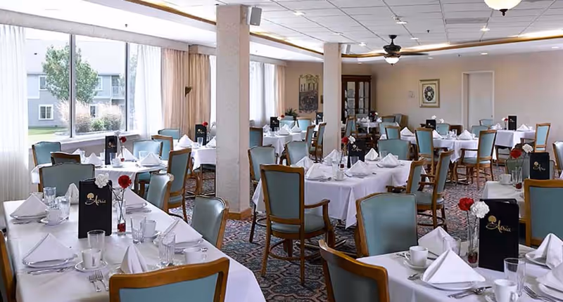 Bright dining room with neatly set tables and upholstered chairs, white tablecloths and folded napkins, and large windows along one side.