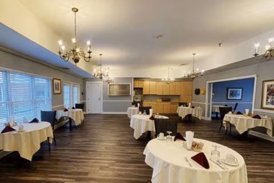 A dining room with several round tables covered with white tablecloths, each set with cups, glasses, silverware, and folded burgundy napkins. The room has wooden flooring, multiple chandeliers hanging from the ceiling, large windows with blinds on the left side, and a kitchenette area with wooden cabinets at the back.
