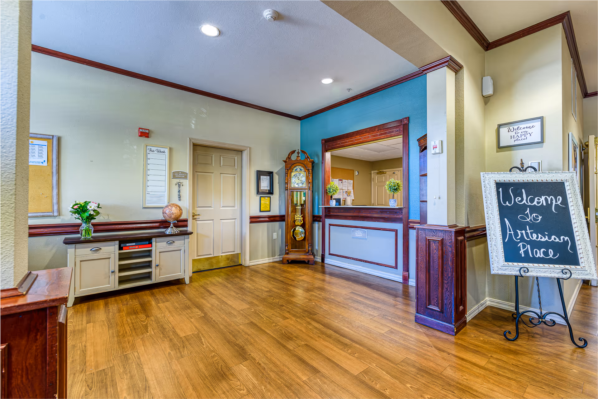 Interior view of a reception area in an assisted living facility with wooden flooring, a small cabinet with a globe and flowers, a grandfather clock, and a reception window with two small potted plants. A chalkboard sign on an easel reads 'Welcome to Artesian Place'.