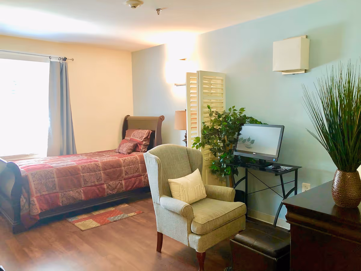 A cozy bedroom with a single bed covered in a patterned quilt, a cushioned armchair with a pillow, a small desk with a computer monitor, and decorative plants. The room has light blue and beige walls and wooden flooring.