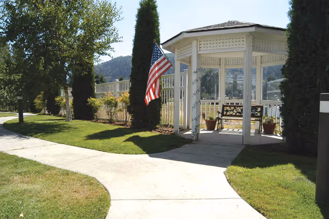 A white wooden gazebo with a bench inside, surrounded by green grass and trees. An American flag is mounted on a tree near the gazebo. A curved concrete pathway leads to the gazebo, with a white fence and hills visible in the background under a clear sky.