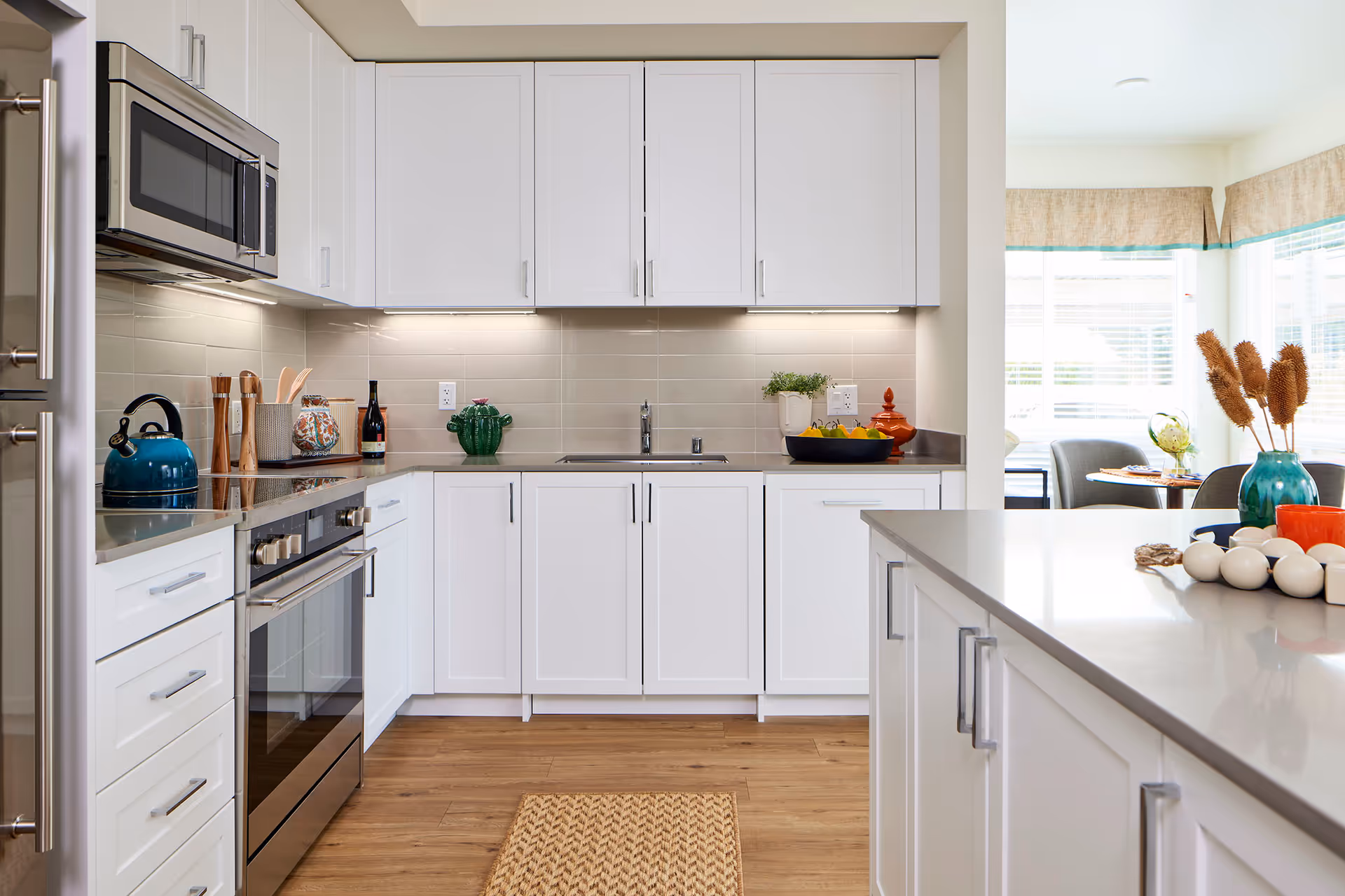 A modern kitchen with white cabinets, stainless steel appliances including a microwave and oven, a blue kettle on the stove, and various decorative items on the countertops. The kitchen has a light wood floor and a beige tiled backsplash. In the background, there is a dining area with a round table and chairs near large windows with blinds.