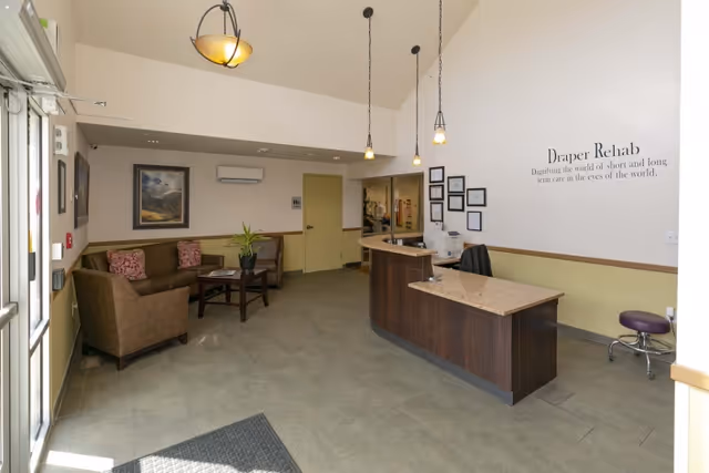 Reception area of Draper Rehabilitation and Care Center with a curved wooden reception desk, three hanging pendant lights, a seating area with brown sofas and a coffee table, framed artwork on the walls, and a wall sign that reads 'Draper Rehab Dignifying the world of short and long term care in the eyes of the world.'