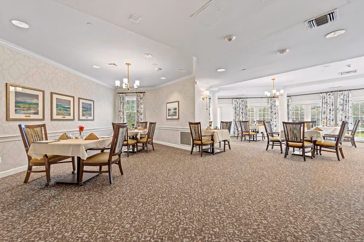 Bright, elegant dining room with multiple round and square tables set with tablecloths and chairs, patterned carpet, framed artwork, and large windows.