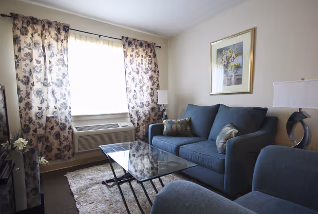 A cozy living room with a blue sofa and matching armchair, a glass coffee table, a beige rug, floral curtains on a window with an air conditioning unit below it, a framed floral painting on the wall, and two modern table lamps on either side of the sofa.