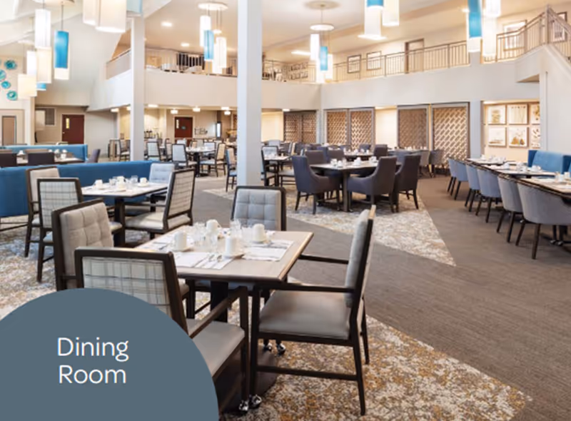 Spacious, well-lit dining room with multiple tables and chairs set for meals in a senior living facility.