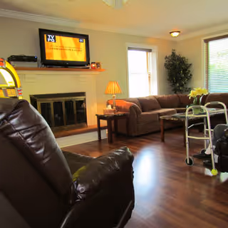 A cozy living room with a brown leather recliner in the foreground, a brown sofa against the wall, a wooden coffee table, a side table with a lamp, a fireplace with a TV mounted above it, and a walker near the sofa. There are windows with blinds and a potted plant in the corner.