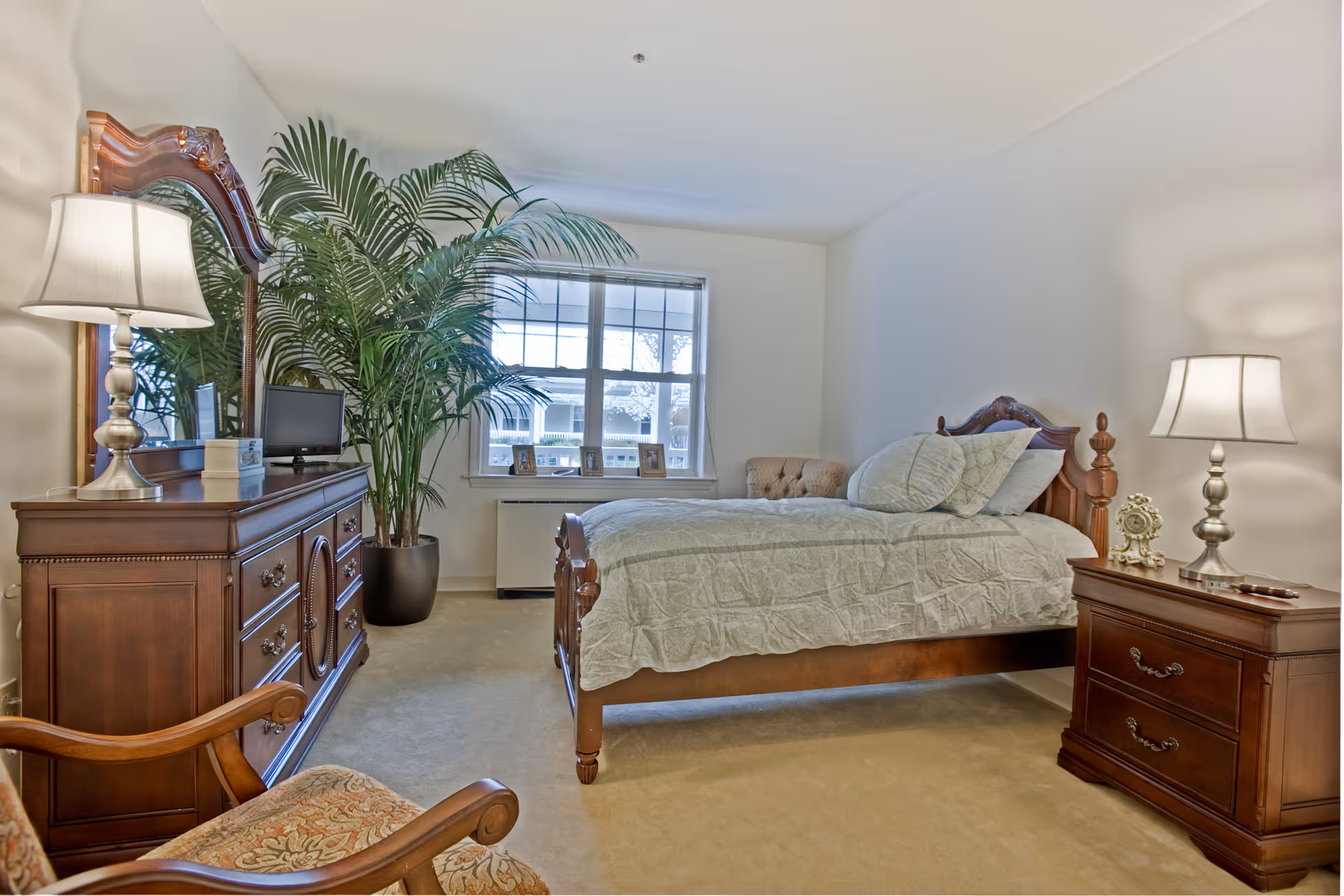 A well-lit bedroom featuring a wooden bed with light-colored bedding and pillows, a wooden nightstand with a lamp and decorative clock, a wooden dresser with a mirror and a small TV, a large potted plant near the window, and a cushioned armchair in the foreground.