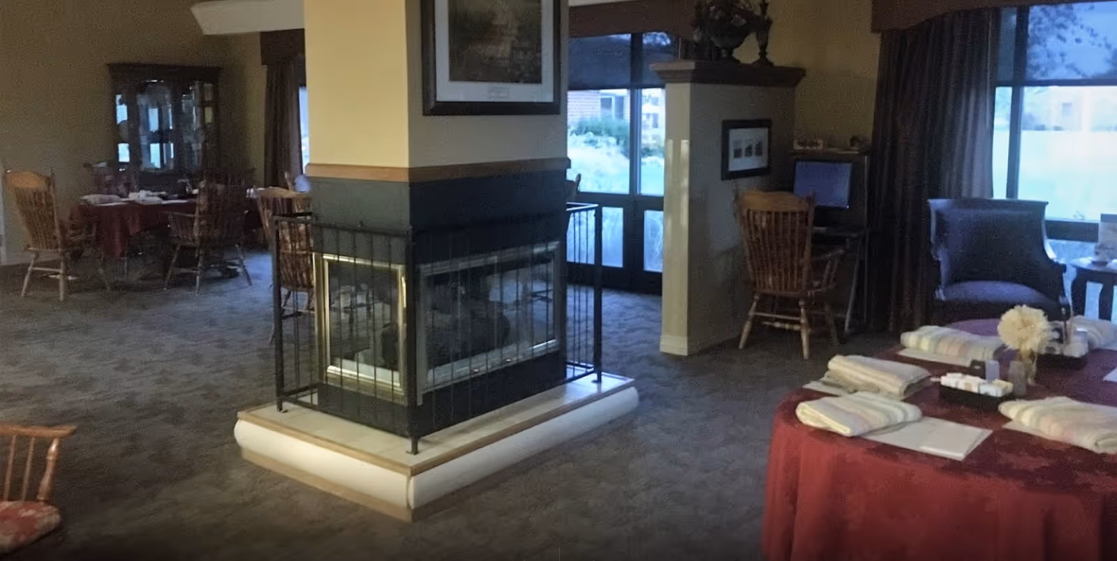 Communal senior living room with a central glass-enclosed fireplace, seating areas, dining tables, and a table set with folded linens.