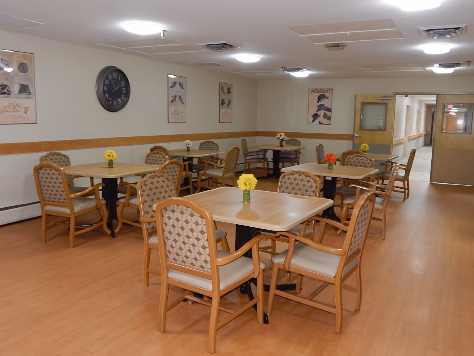 A dining room with multiple square wooden tables and cushioned chairs arranged neatly. Each table has a small vase with colorful flowers. The room has light-colored walls with framed artwork and a large clock. There is a hallway visible through an open door at the back.