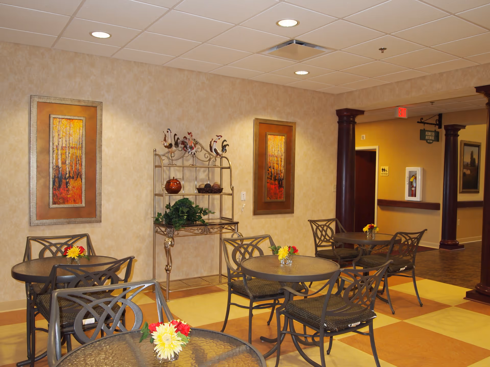 Indoor seating area with round glass tables and metal chairs, each table decorated with a small vase of flowers. The walls are beige with two framed paintings of autumn trees. A decorative metal shelf with ornamental roosters and plants is against the wall. The floor has a checkered pattern in yellow and brown. Dark wooden columns and a hallway with a restroom sign and exit sign are visible in the background.
