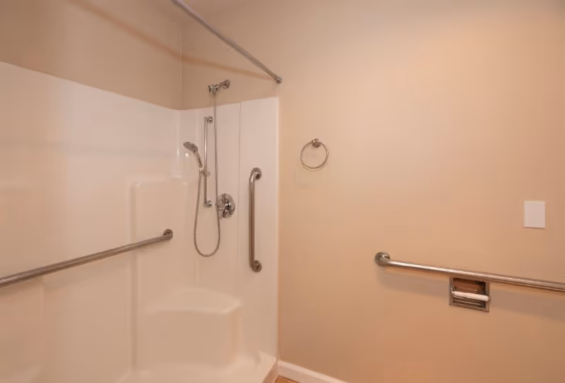A bathroom shower area with beige walls, a built-in white shower seat, a handheld showerhead, and multiple stainless steel grab bars for support. There is also a towel ring and a recessed toilet paper holder on the wall.