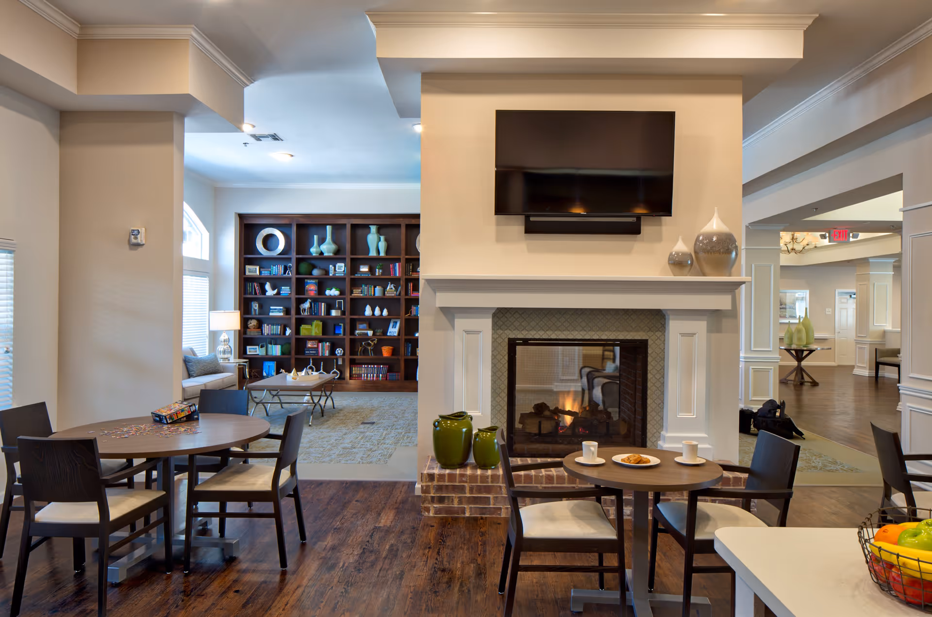 A cozy common area in Twin Rivers Assisted Living and Memory Care featuring a lit fireplace with a TV mounted above it. There are two small round tables with chairs around them, one with a plate of cookies and cups. In the background, there is a large bookshelf filled with decorative items and books, and a seating area with a window letting in natural light. The room has warm wooden flooring and neutral-colored walls.