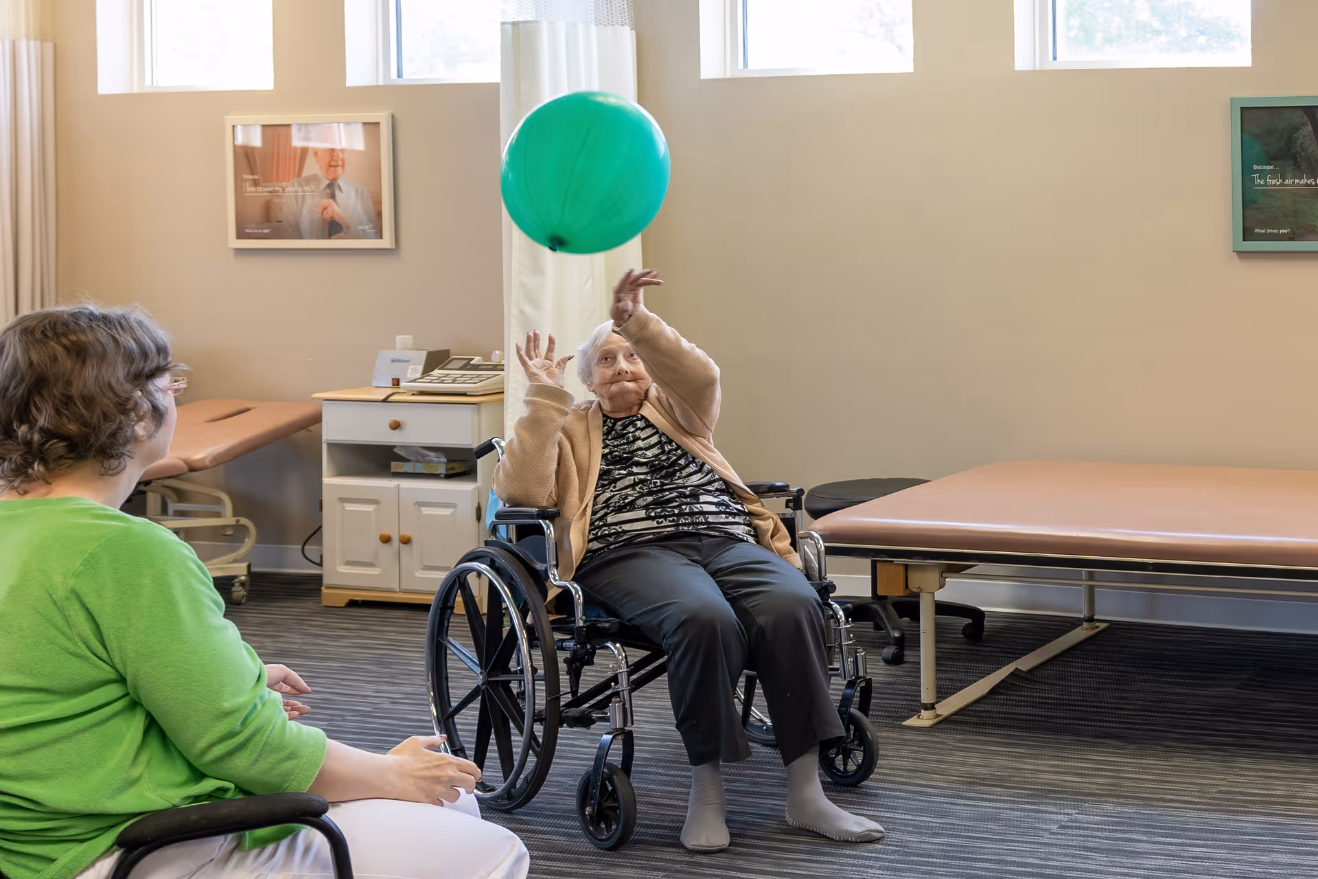 An elderly woman in a wheelchair playing with a green balloon inside a rehabilitation room. Another person in a green shirt is sitting nearby, watching her. The room has medical examination tables, a cabinet, and framed pictures on the walls.