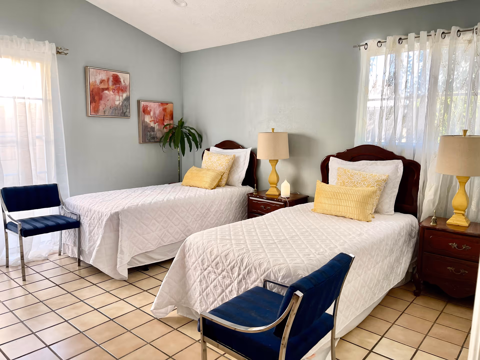 A bright bedroom with two twin beds dressed in white quilts, yellow pillows, bedside tables with lamps, and blue chairs on a tiled floor.